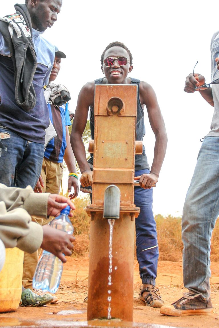 A young man using a water pump surrounded by friends in a rural area.